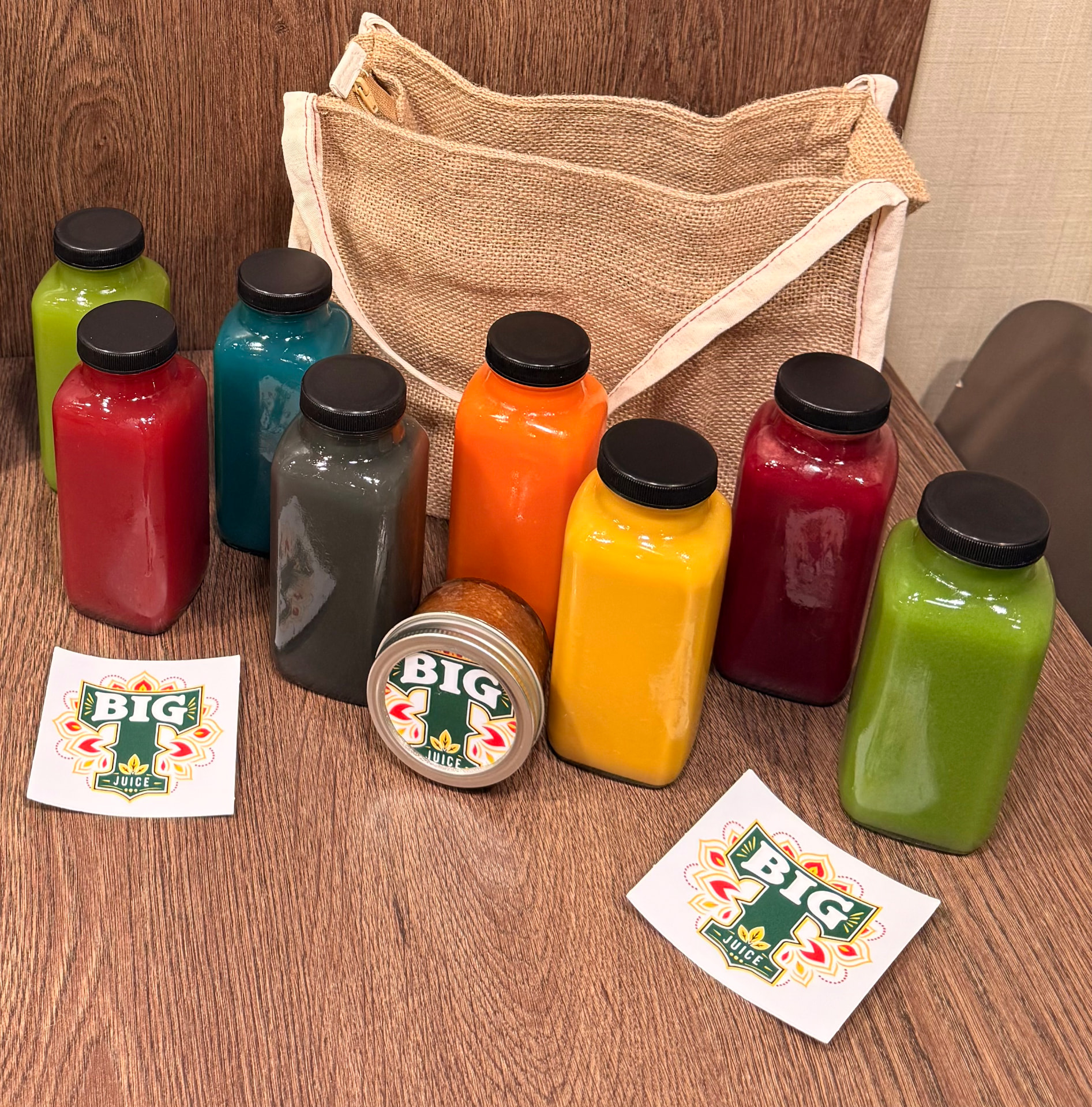 A colorful lineup of BIG T Juice bottles beside the official logo and jute tote